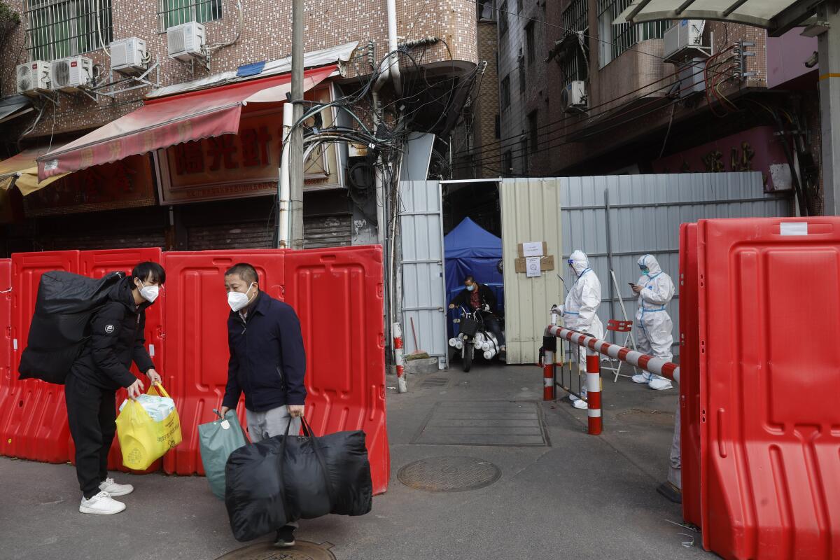 Migrant workers with their belongings leave a barricaded village while people in protective suits stand nearby.