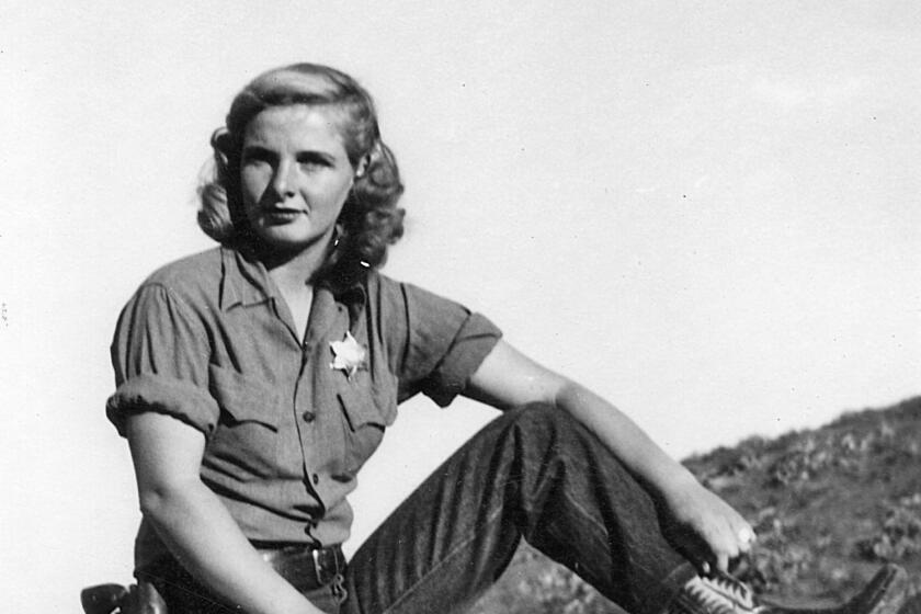 A black-and-white frame of a woman wearing cowboy boots, jeans, belt with a pistol and a sheriff's star, sitting on a beam. 