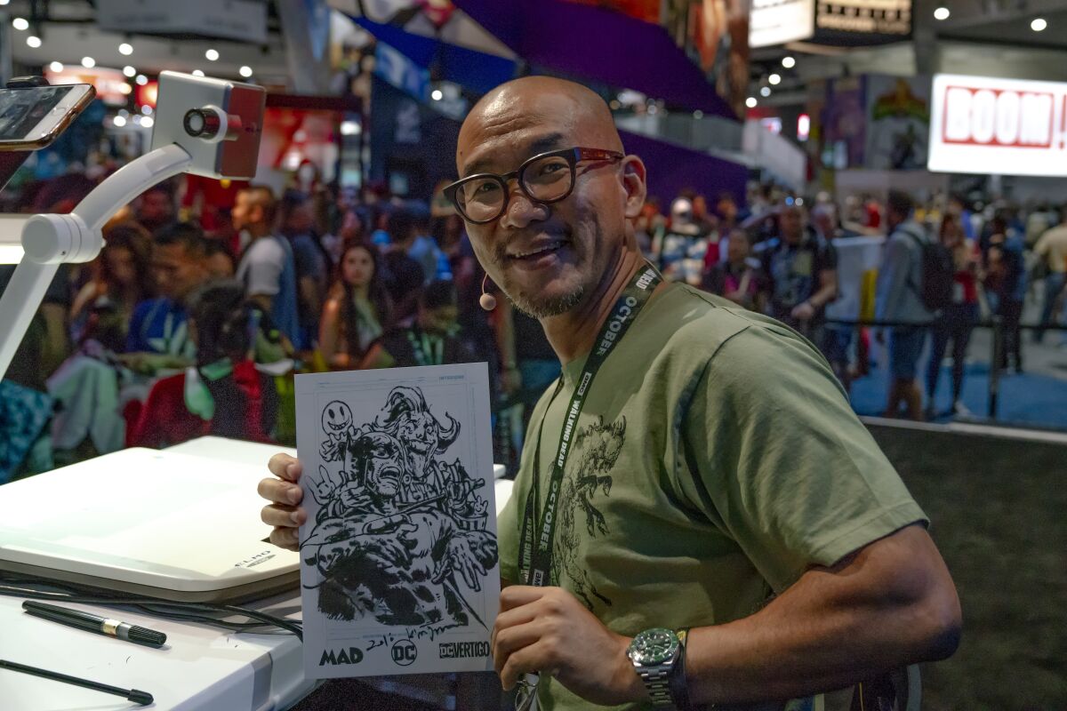 A bald man wearing glasses and smiling while holding up a drawing in front of a crowd.