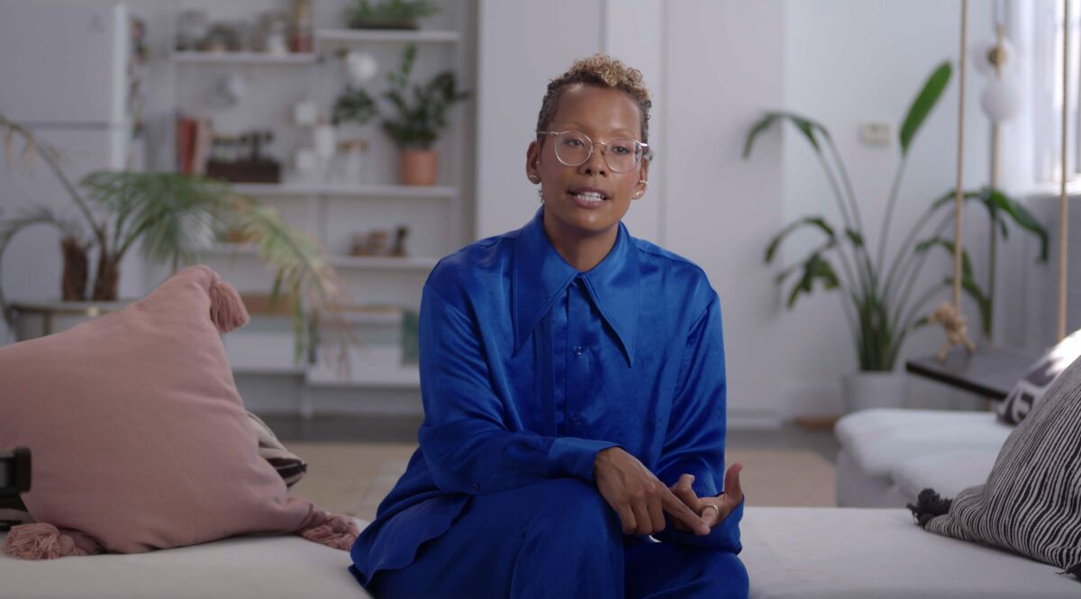 A woman in a blue blouse speaks while sitting on a white couch.