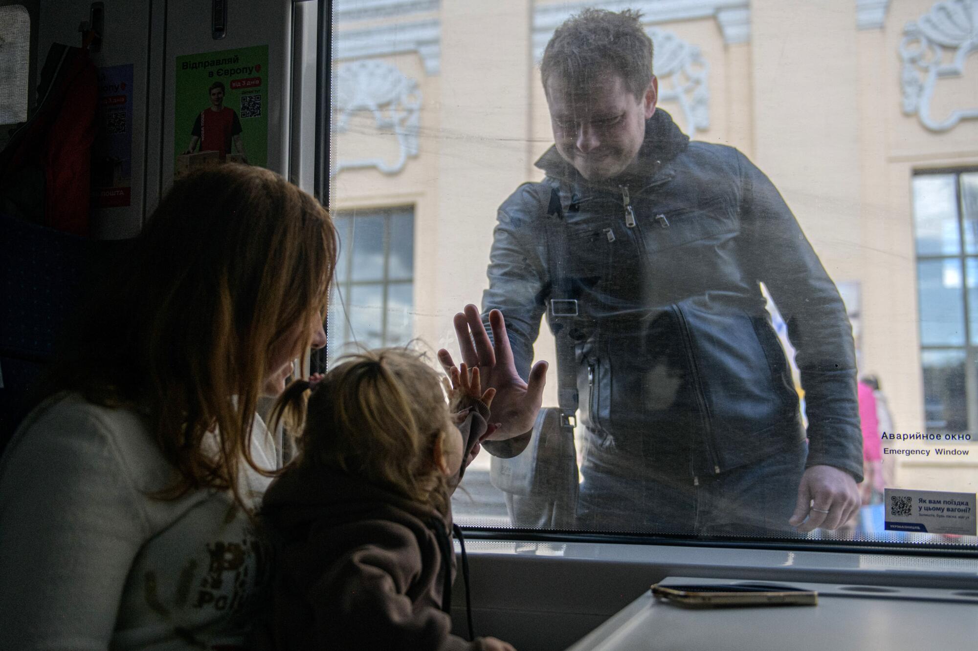 A man holds his palm to the glass window separating him from a woman and a child, who puts her palm to his 