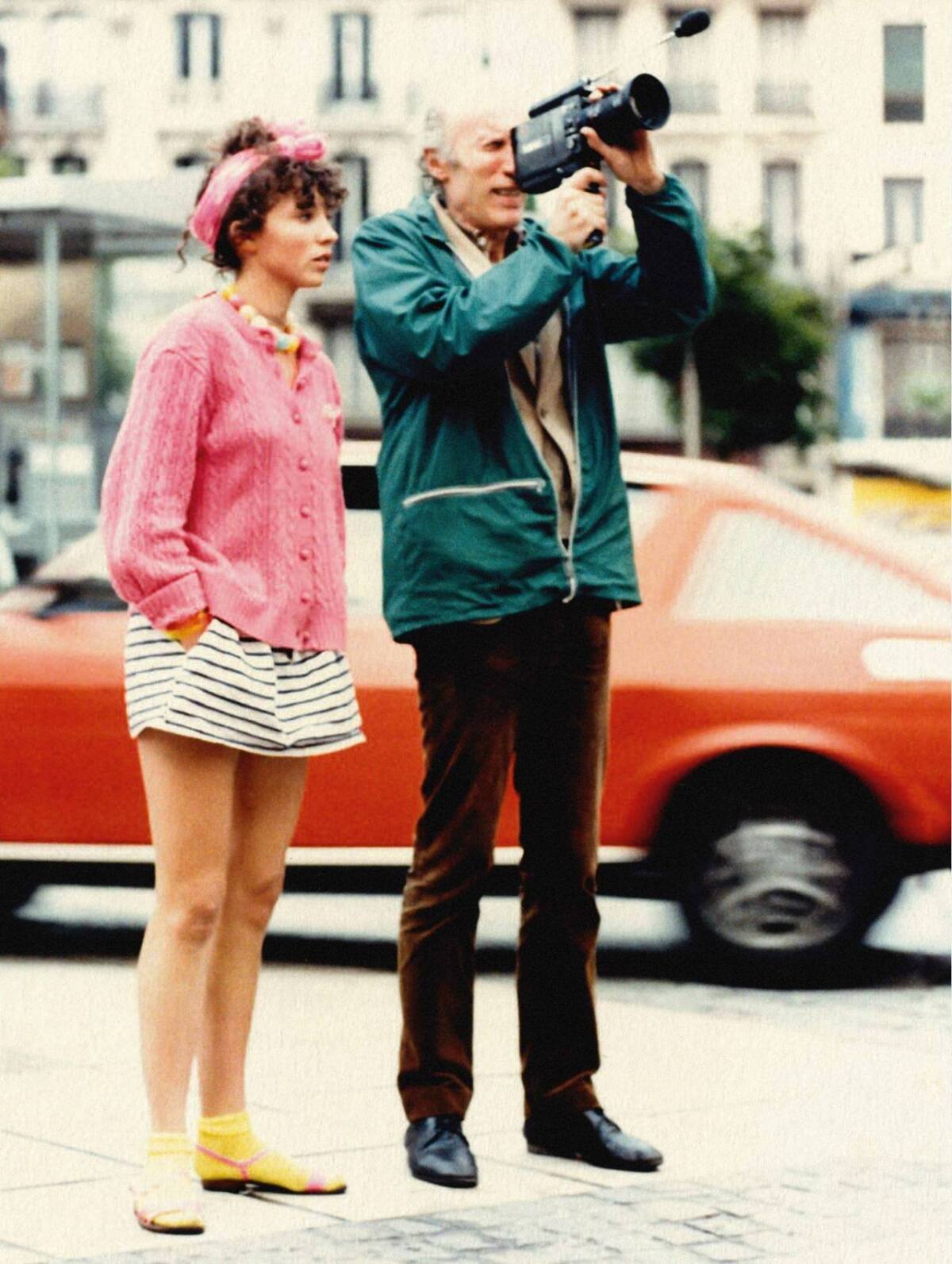 A woman in a pink jacket stands with a man shooting a camera.