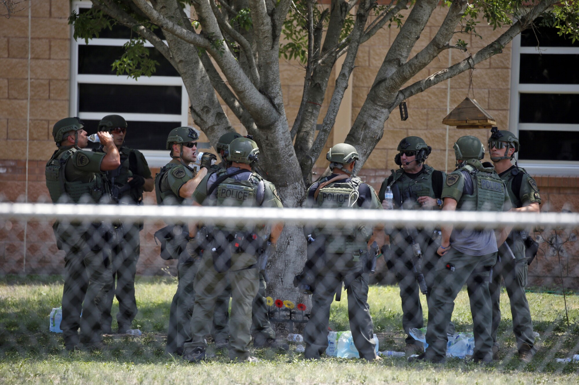 A group of officers in police uniform, some drinking water, stand under a tree near a building with windows
