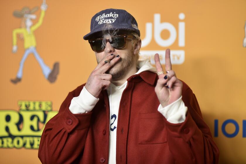 A man in a baseball cap and sunglasses smokes a cigarette and flashes a peace sign