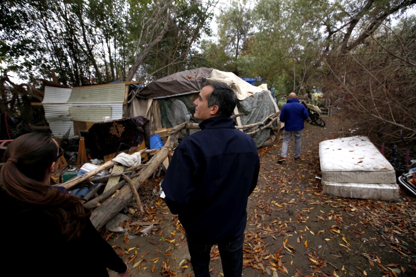 Eric Garcetti stands in front of a tent and a structure made of wood and corrugated metal in a woodsy area