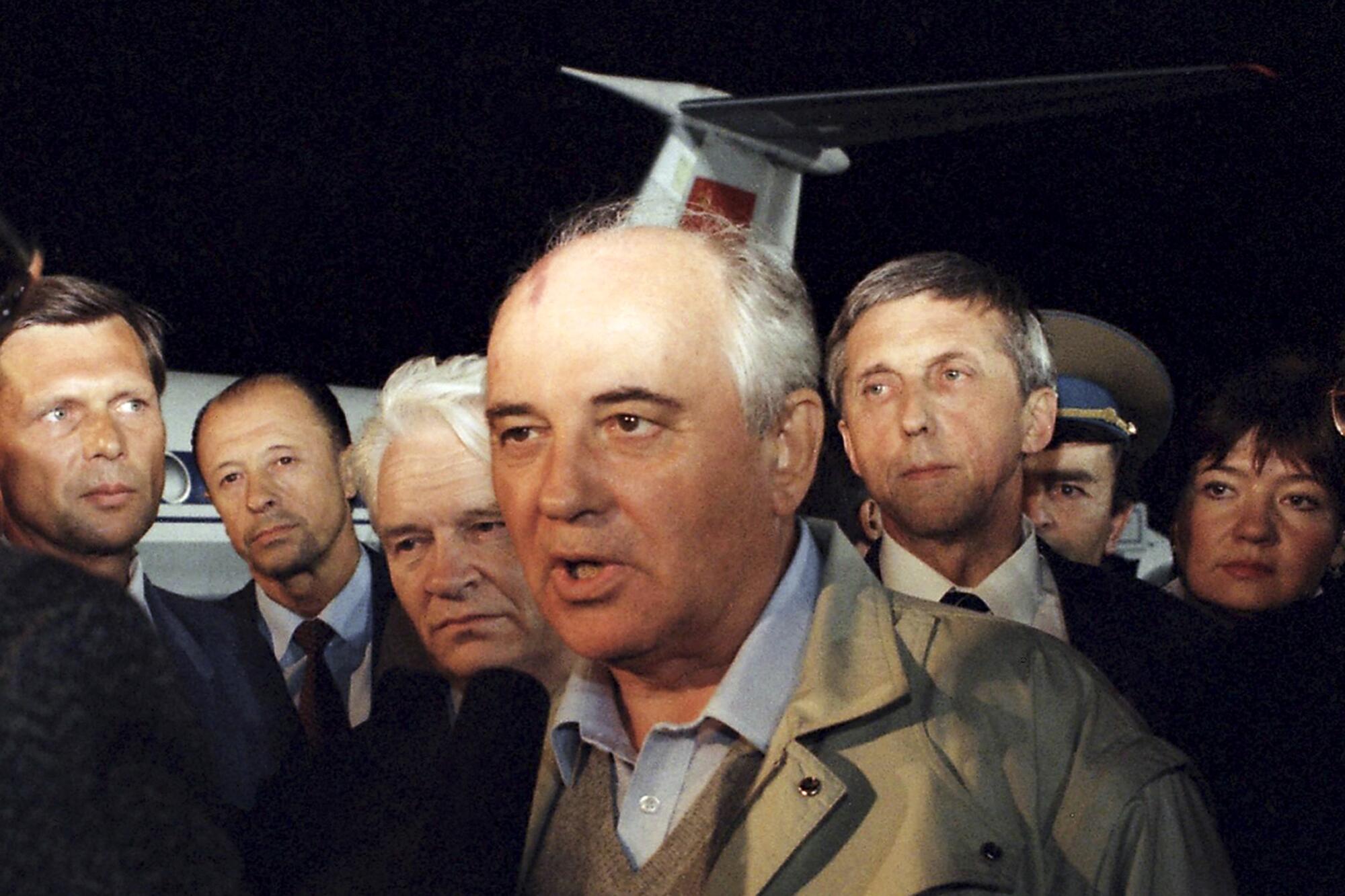 A man in a light blue shirt and khaki jacket speaks as he is surrounded by other people near a plane 