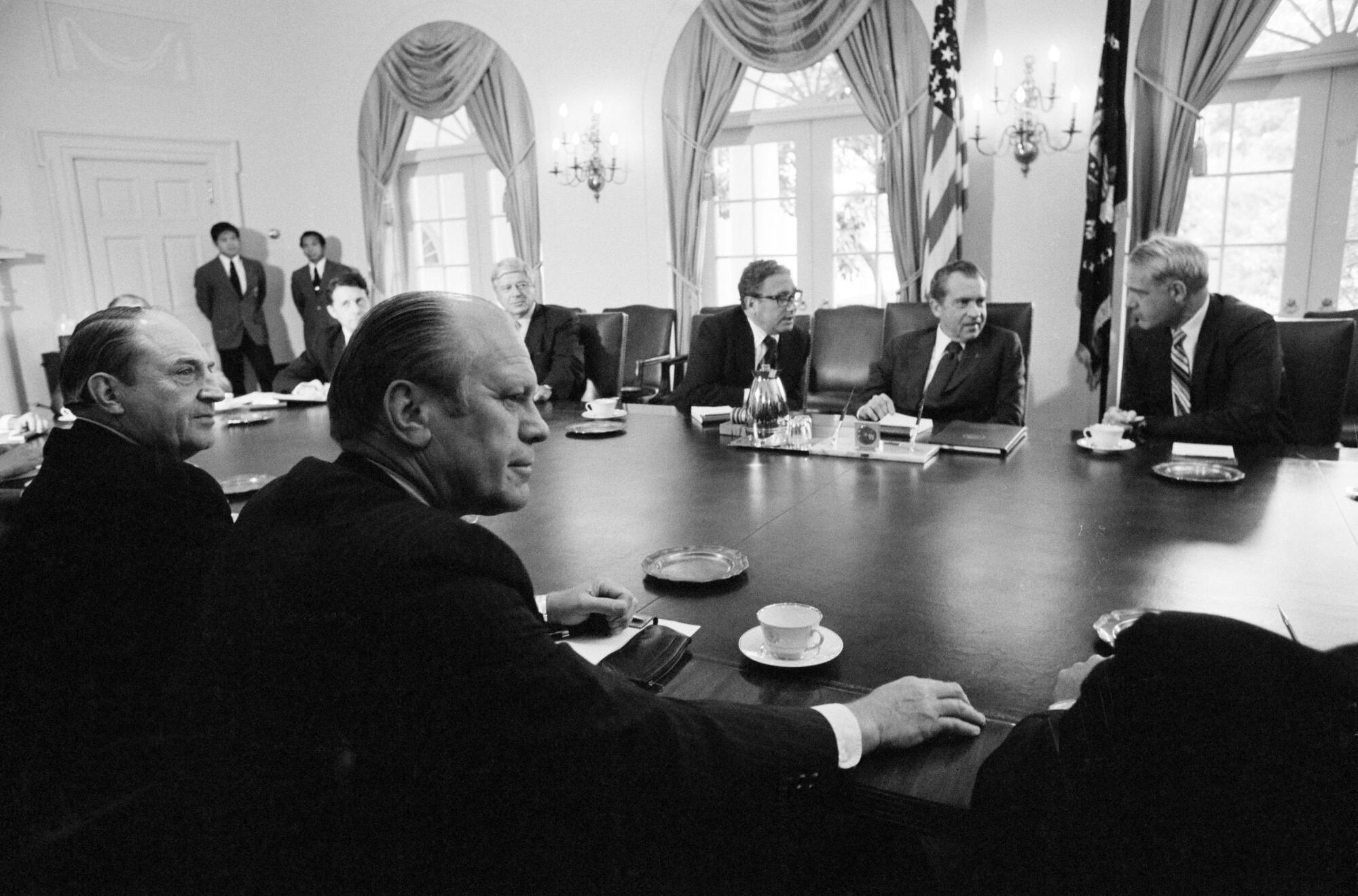Uma foto em preto e branco de autoridades sentadas em uma mesa em uma sala ornamentada, Gerald Ford em primeiro plano, em frente a Richard Nixon