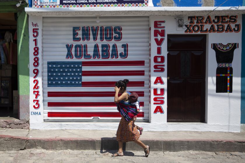 In this April 3, 2020 photo, a woman carrying a child walks past a closed courier business featuring a U.S. flag and the Spanish phrase: "Send to U.S.A" in the largely indigenous town of Joyabaj, Guatemala, where half of the residents depend on remittances, almost all from the U.S. The devastation wrought by COVID-19 across the developed world is cutting into the financial lifelines for people across Latin America, Africa and Asia. (AP Photo/Moises Castillo)