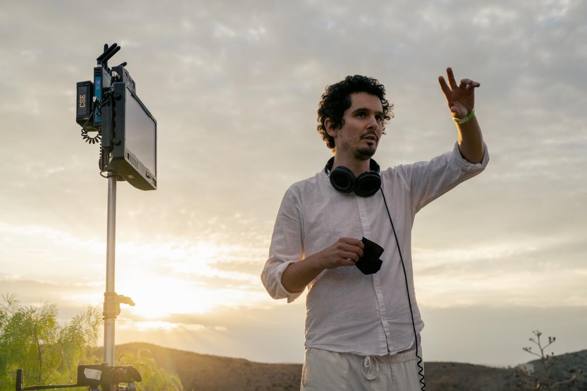 A man dressed in white, with headphones around his neck, stands outside with his arm raised.