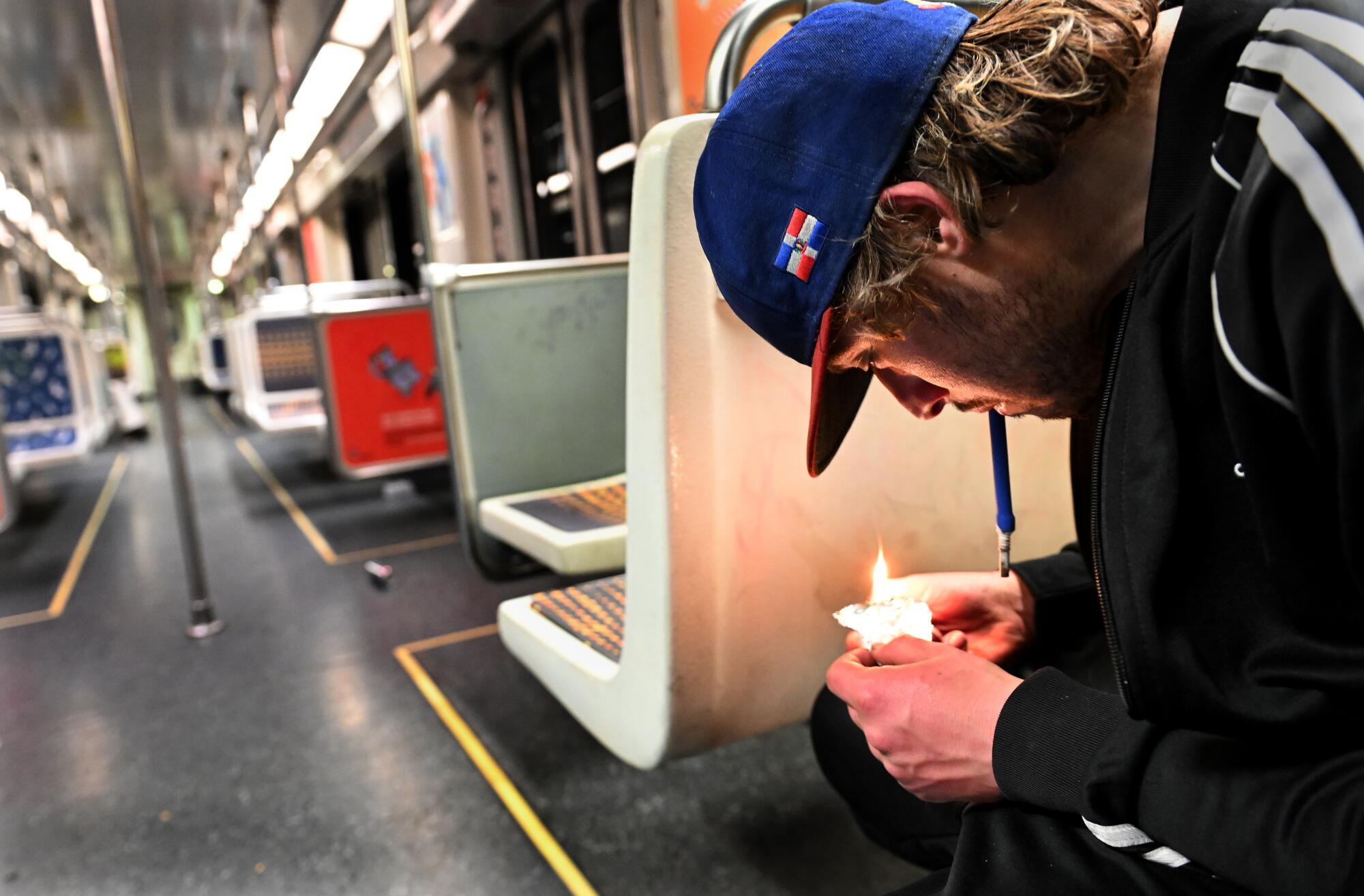 At right, seated on a subway train, a young man with dark blond hair and a baseball cap smokes fentanyl.
