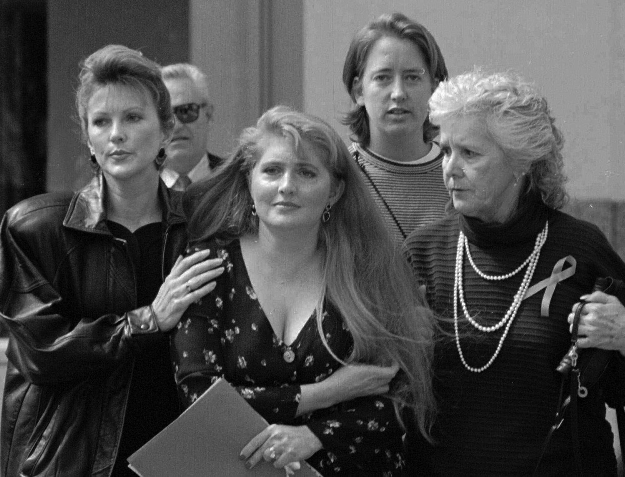 A black and white photo of three friends comforting Eileen Franklin-Lipsker as they walk down a hallway, a man behind them