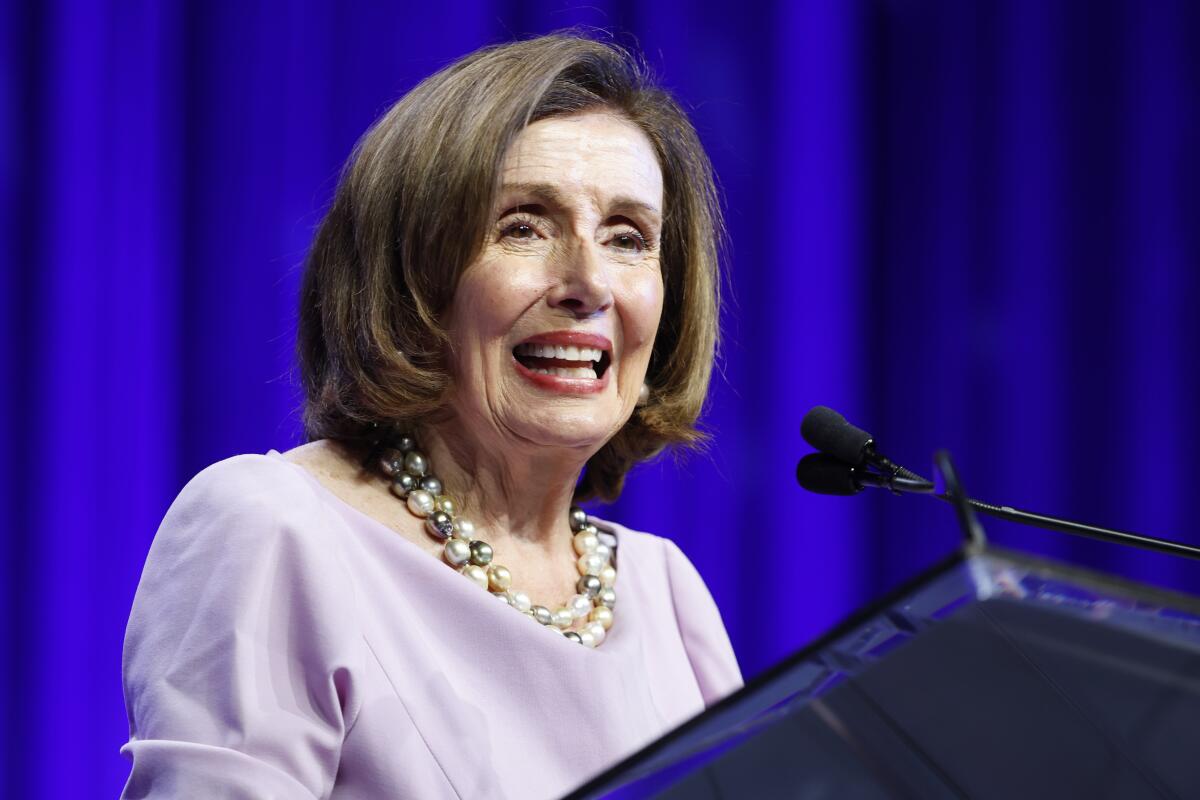 Nancy Pelosi pictured from the shoulders up speaking from a lectern against a royal-blue backdrop