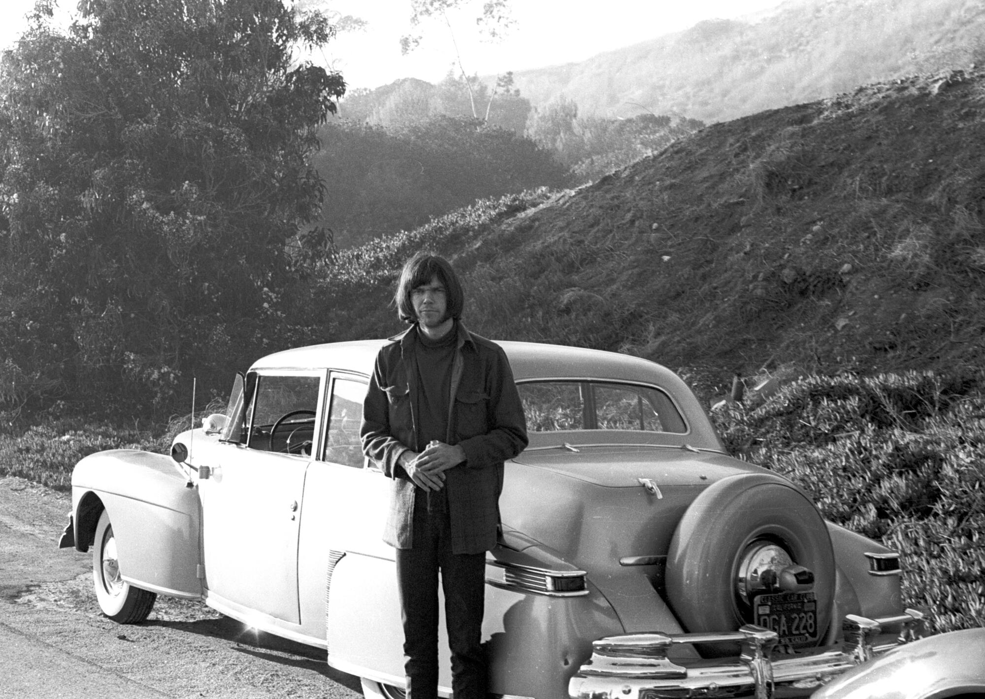 Neil Young in 1967 standing in front of a vintage car amid mountains