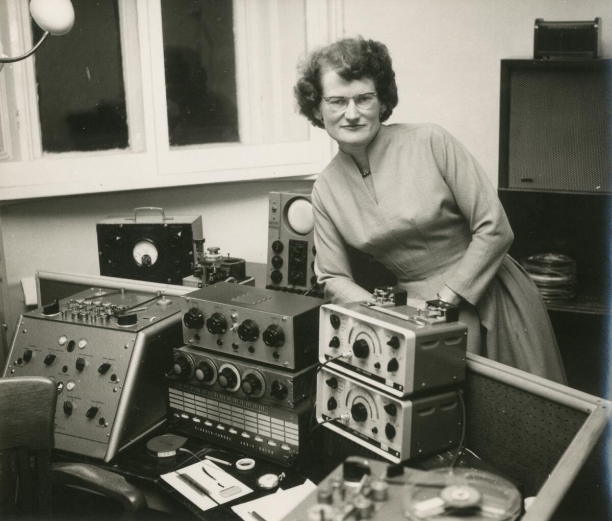 A woman stands beside a bank of electronic equipment.