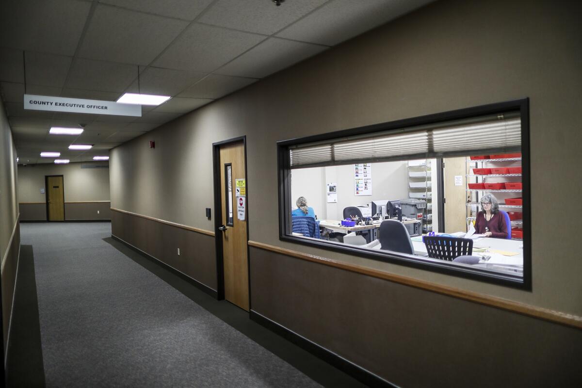 Workers at tables in an office are seen through a large window in a hallway