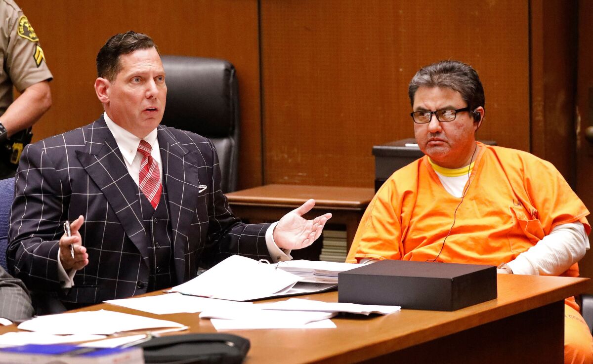 A man talks at a table as another man, in orange, watches in a courtroom.