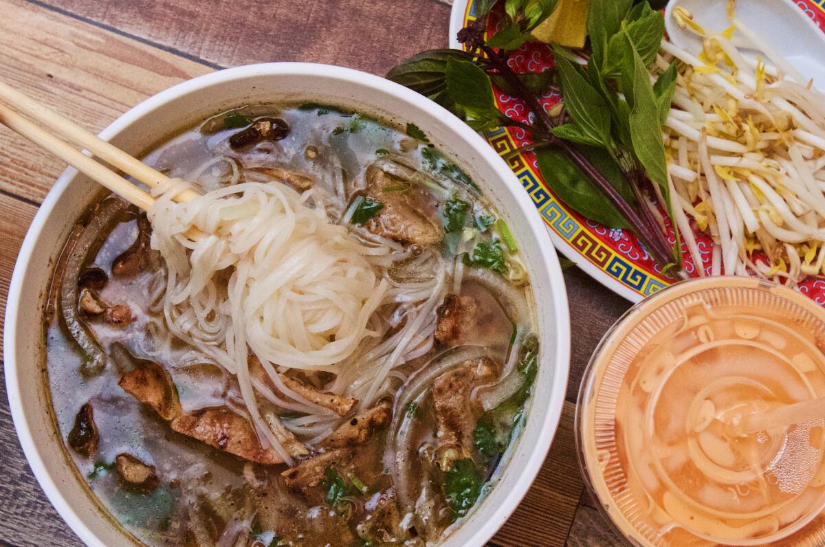 Overhead photo of chopsticks lifting noodles from charred-pork pho near Thai iced tea and a plate of basil and bean sprouts.