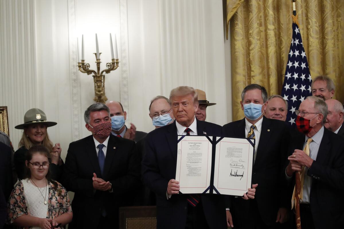 President Trump holds an open folder showing a bill he signed as he stands in front of a group of people. 