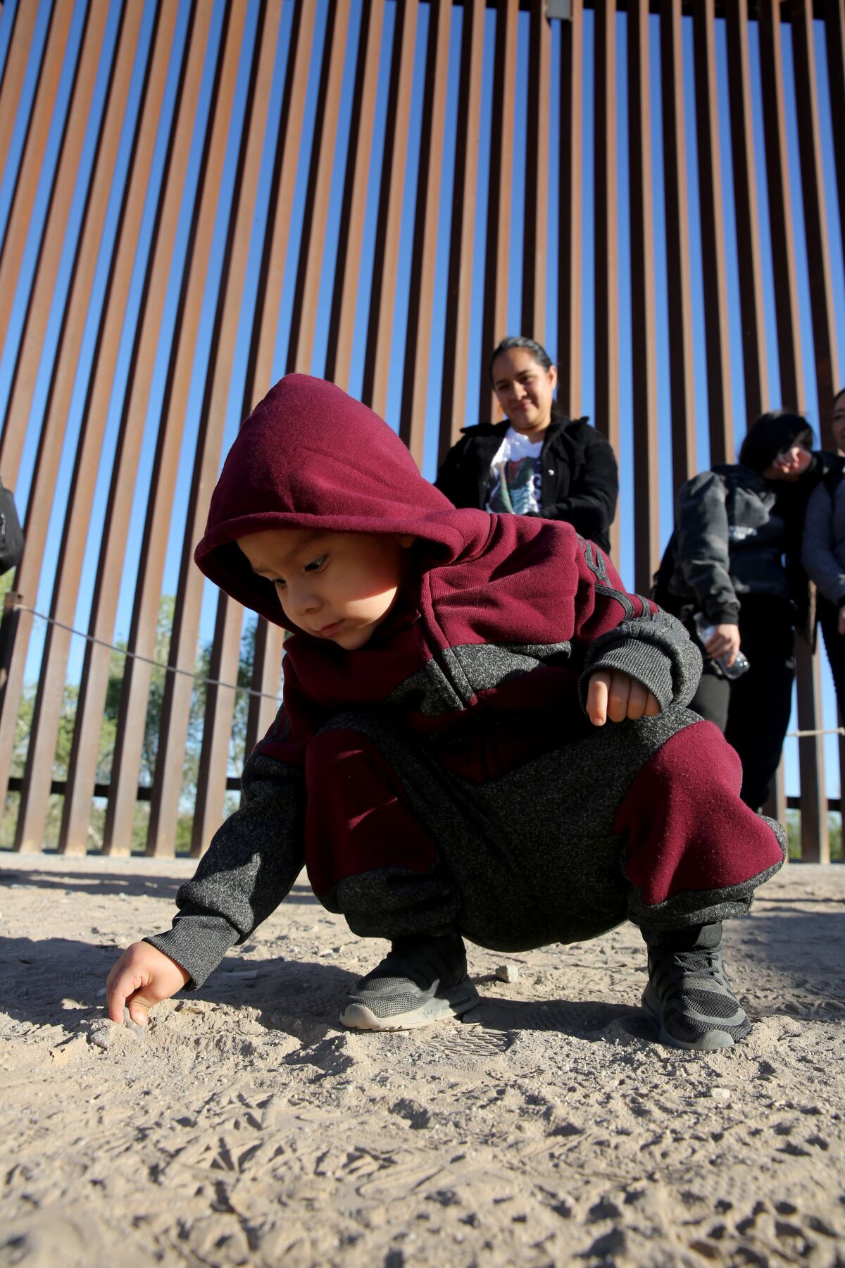 A small boy in a hoodie squatting and drawing on the dusty ground near the border wall as a woman in the background watches