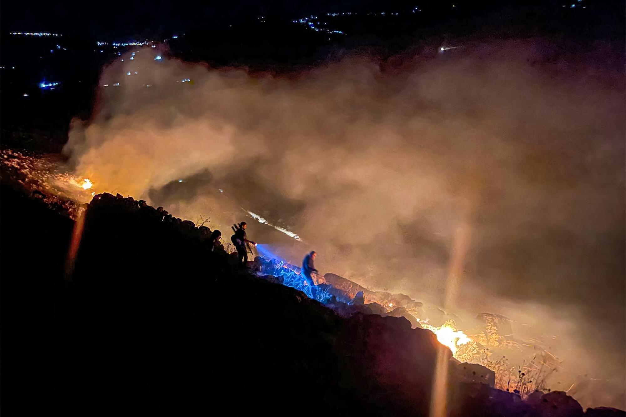 Two people are seen on a dark slope where flames are seen amid rising smoke 