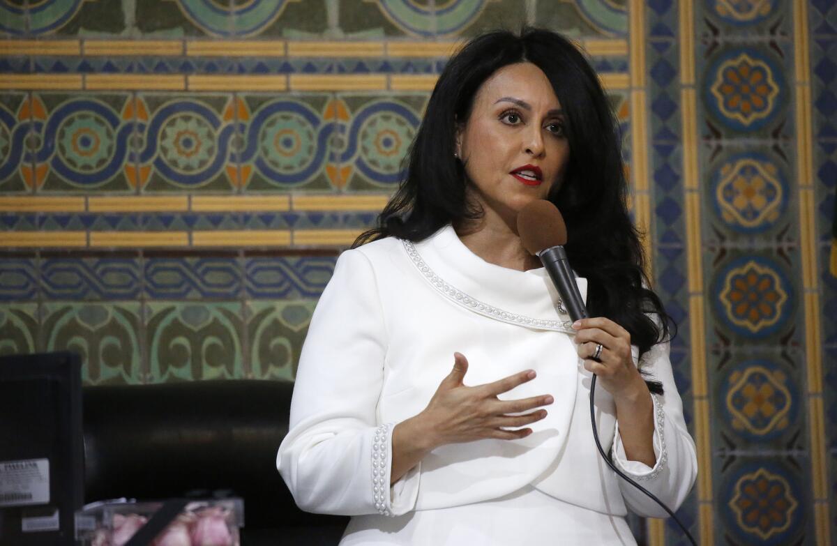 A woman in a white suit stands on a stage speaking into a microphone.