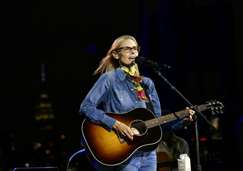 A woman in a denim jacket and pants plays the guitar and sings into a mic
