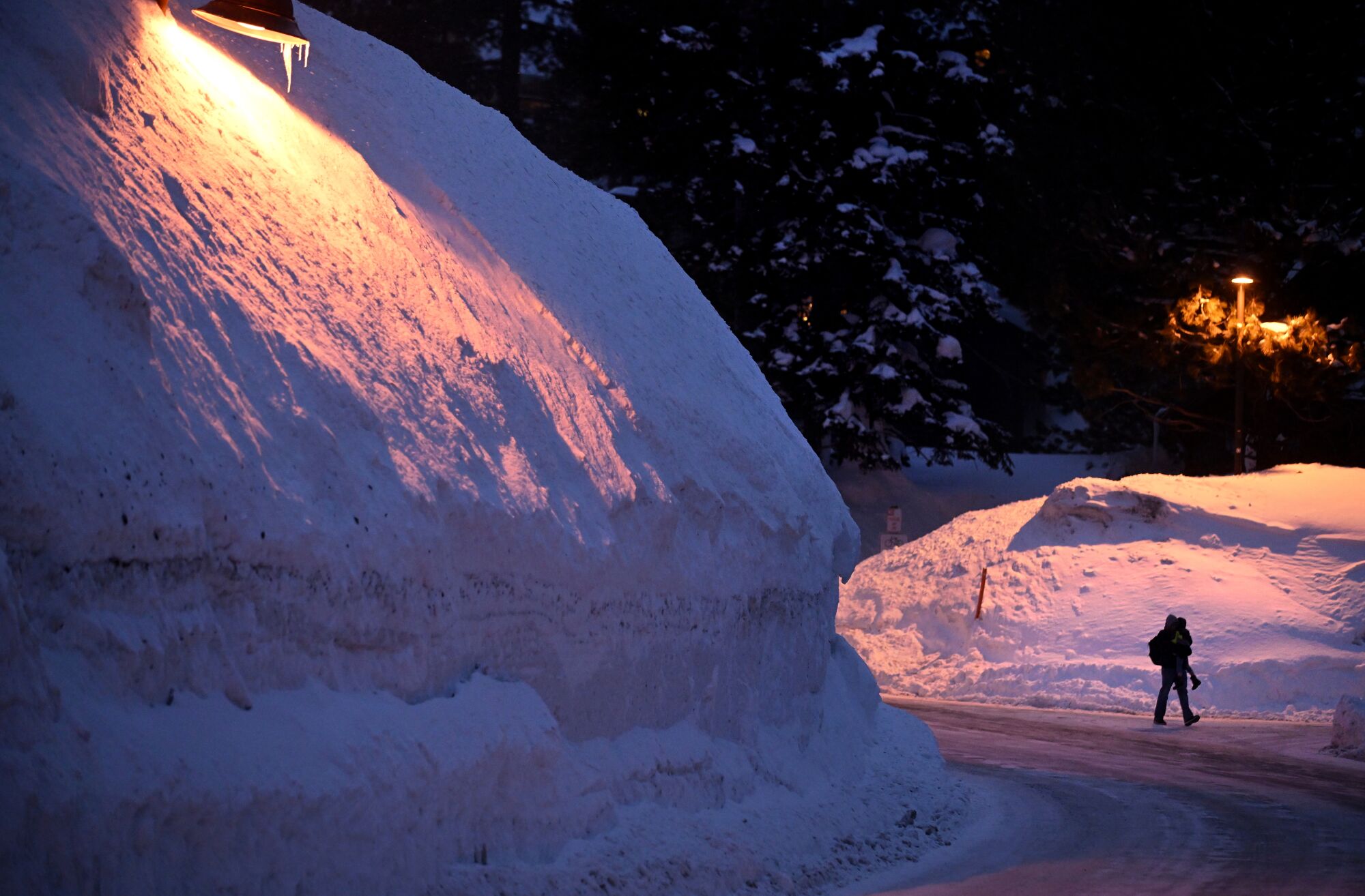 A small figure of a person carrying a child walks at night on a frozen road between huge snow banks lit by street lamps. 