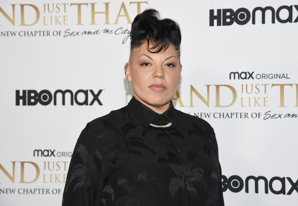 Sara Ramirez standing in a dark formal shirt and a chain necklace in front of a white backdrop with black and gold text