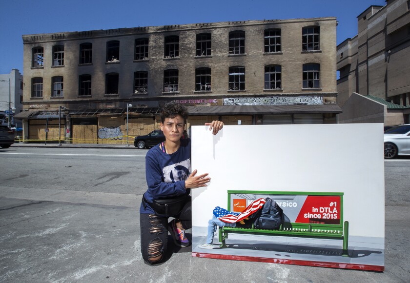 An artist kneels in front of a burnt building with a painting of a man lying on a bench with an American flag draped over him
