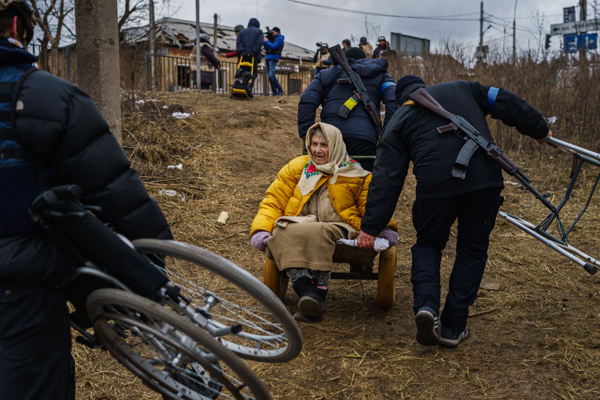 A woman dressed in a warm jacket and scarf is rolled on a cart up a hill by two uniformed people