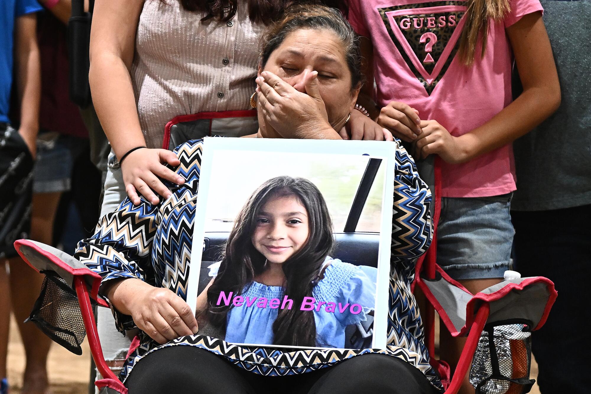 A seated woman with a hand to her face holds a picture of a girl with long dark hair and the name Nevaeh Bravo