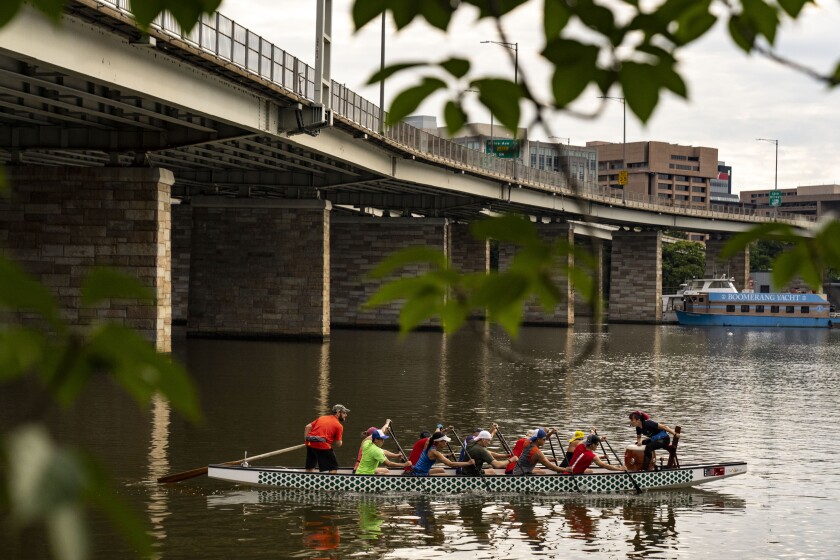People in a long flat boat row in a body of water. In the background is a bridge and some city buildings.