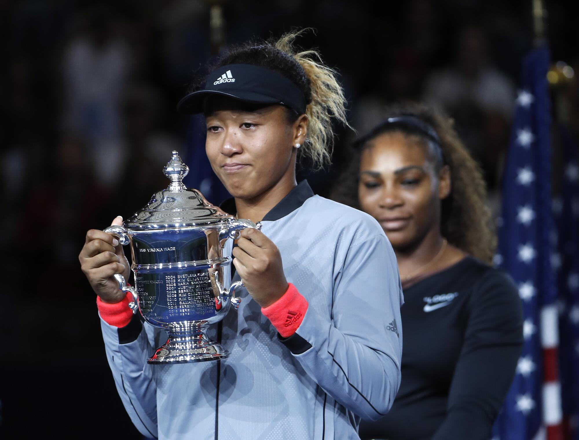 Naomi Osaka holds a large silver trophy with both hands, Serena Williams behind her.