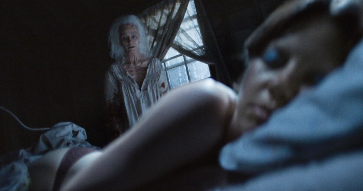 A woman lies on a bed in the horror film "X," with an old woman watching her.
