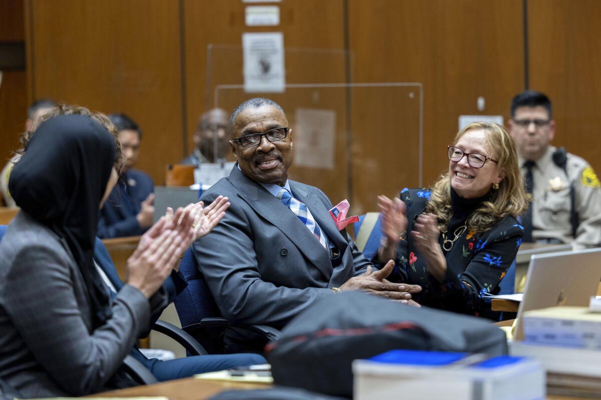 A man sits in court with people sitting on either side of him clapping and smiling as they face him