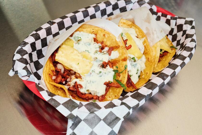 A basket of three adobada tacos topped with pineapple and cream sauce on a metal counter at Cypress Park's Taquería Frontera