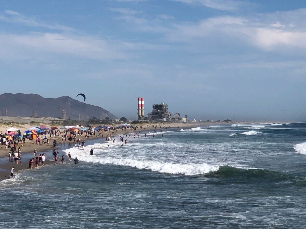 People sit along the shoreline or go into the surf. In the background are mountains and smokestacks.