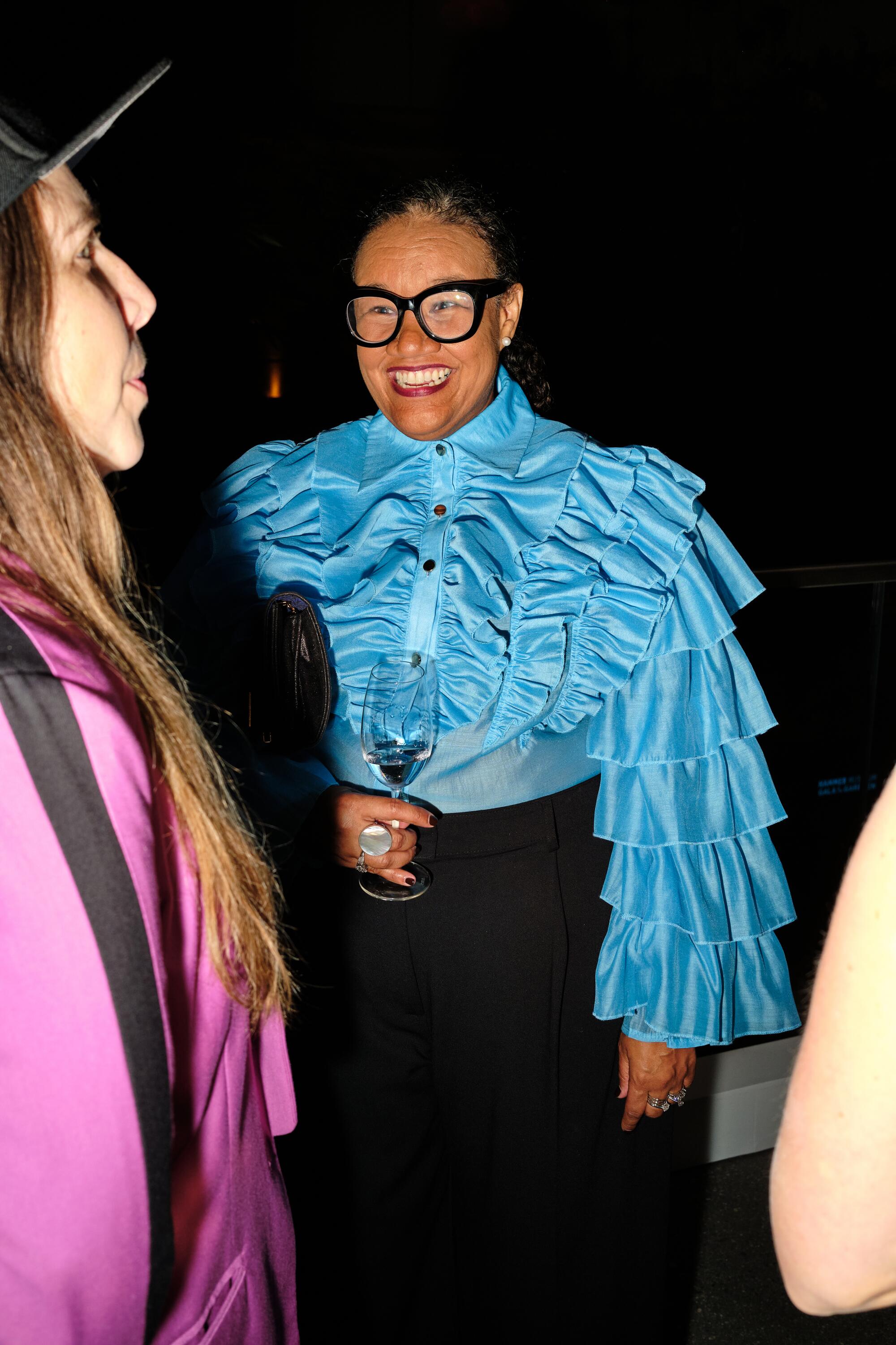 A woman in a bright blue ruffled top and thick black glasses smiles for a picture at the Hammer Museum gala.
