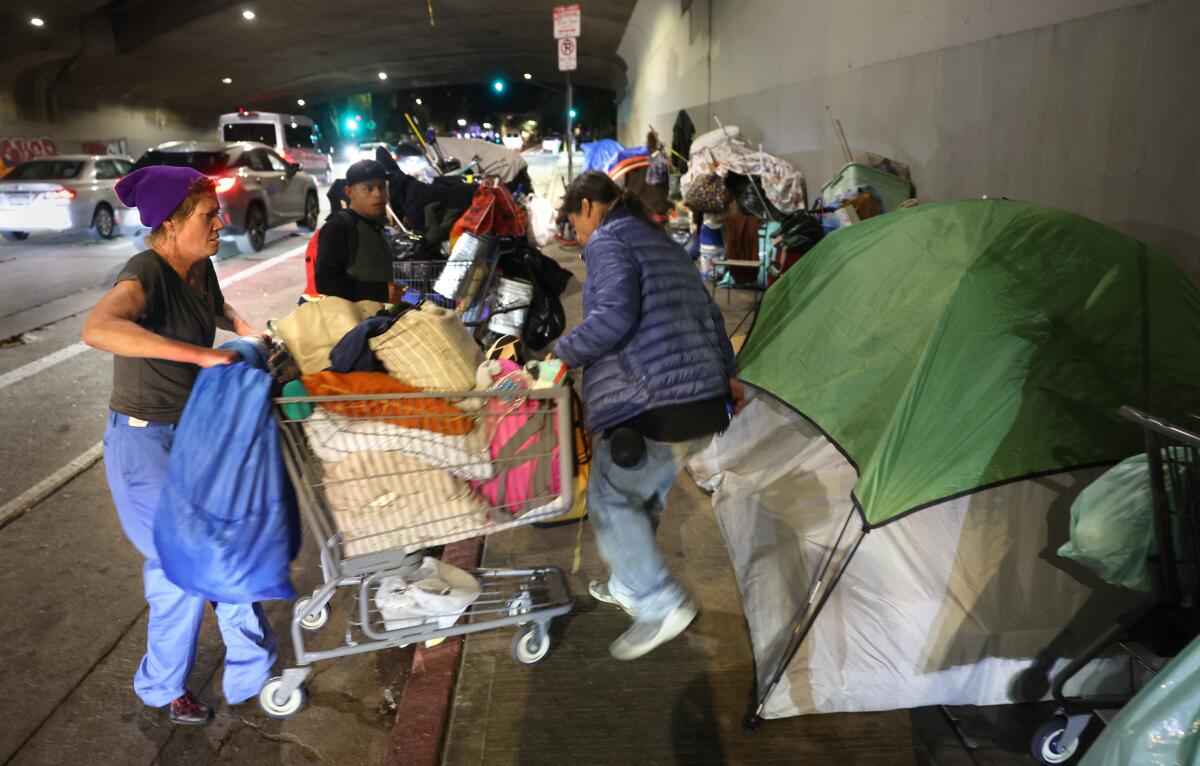 Three people next to a grocery cart and tents at a homeless encampment.
