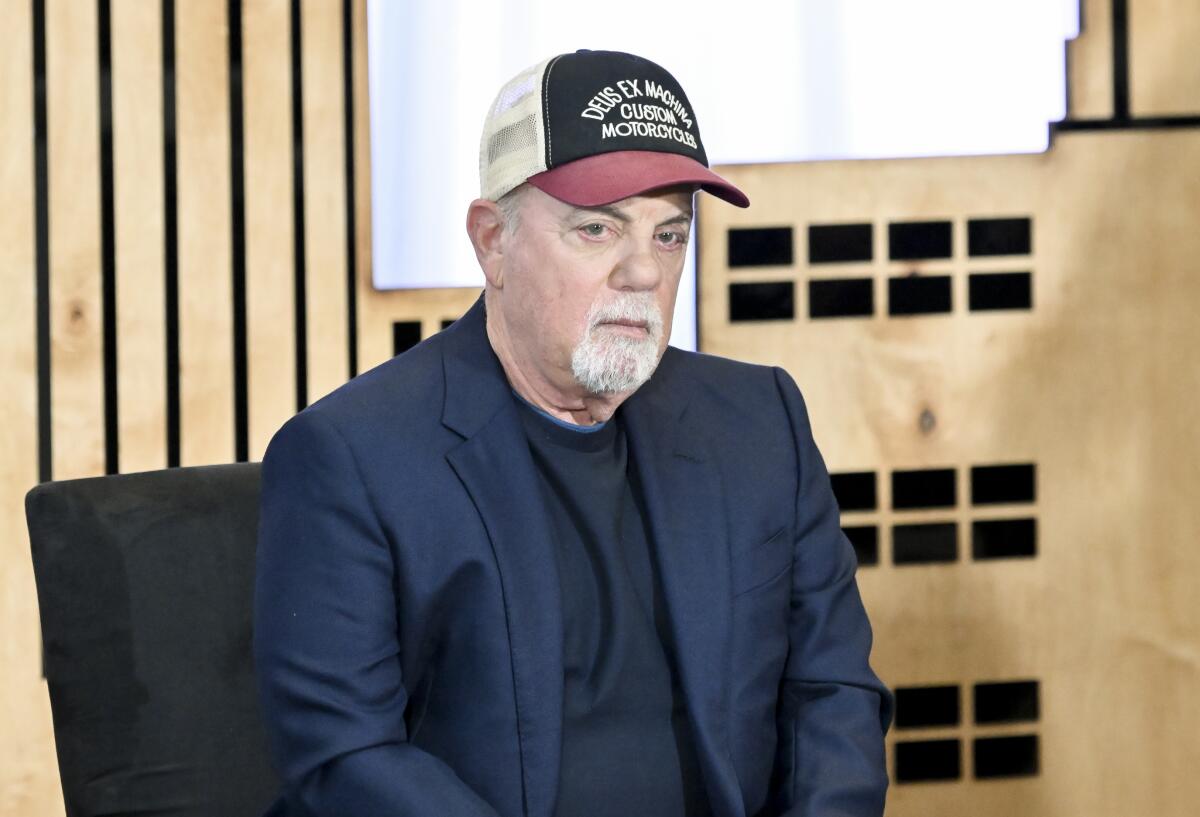 Billy Joel sits in a blue suit jacket and crewneck shirt wearing a trucker cap