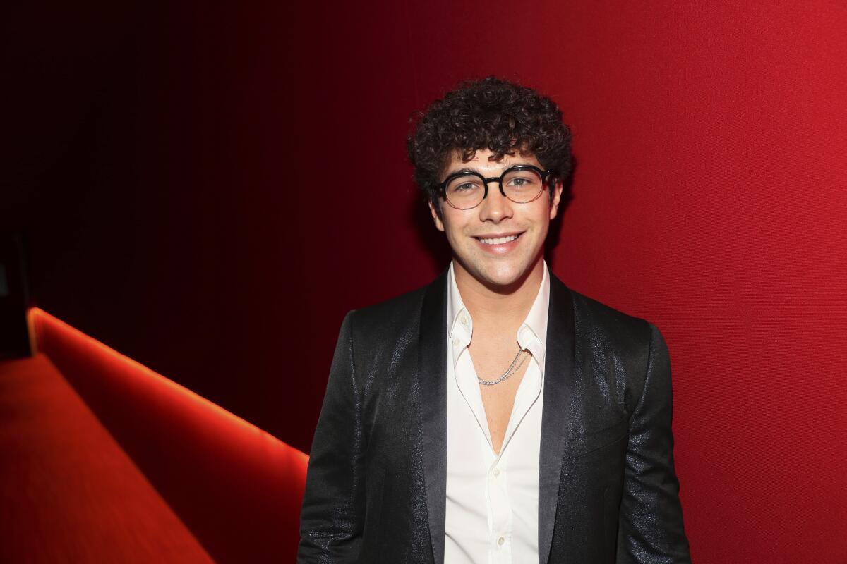 Austin Mahone wearing glasses and showing his curly hair in front of a red backdrop