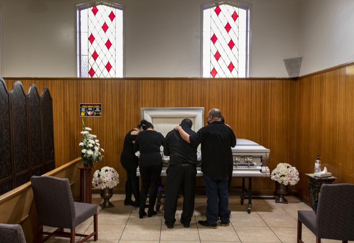 Several people wearing black huddle in front of an open coffin in a funeral home, heads bowed.