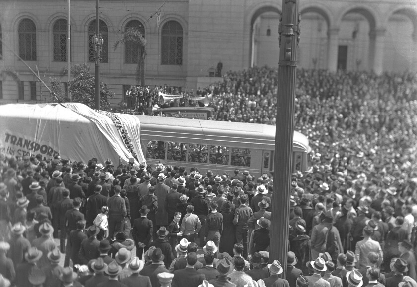 A densely packed crowd, many of them men in hats, surrounds a long trolley that is half covered by a tarp.