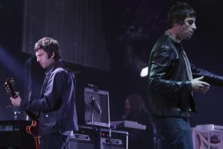 Noel Gallagher playing guitar while facing away from his brother Liam Gallagher onstage surrounded by equipment.