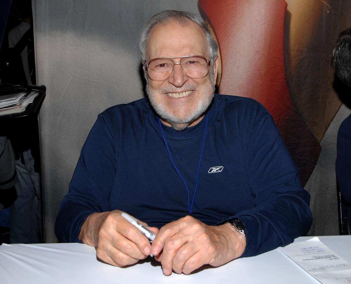 John Romita Sr. sits at a table smiling with a pen in his hand