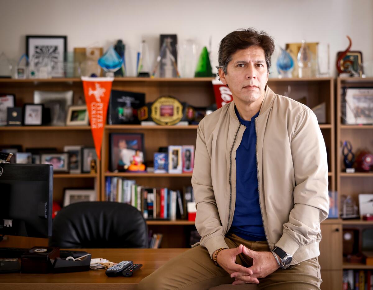A man with dark hair, in a beige jacket and khaki pants, sits on the edge of a desk with bookshelves behind him 