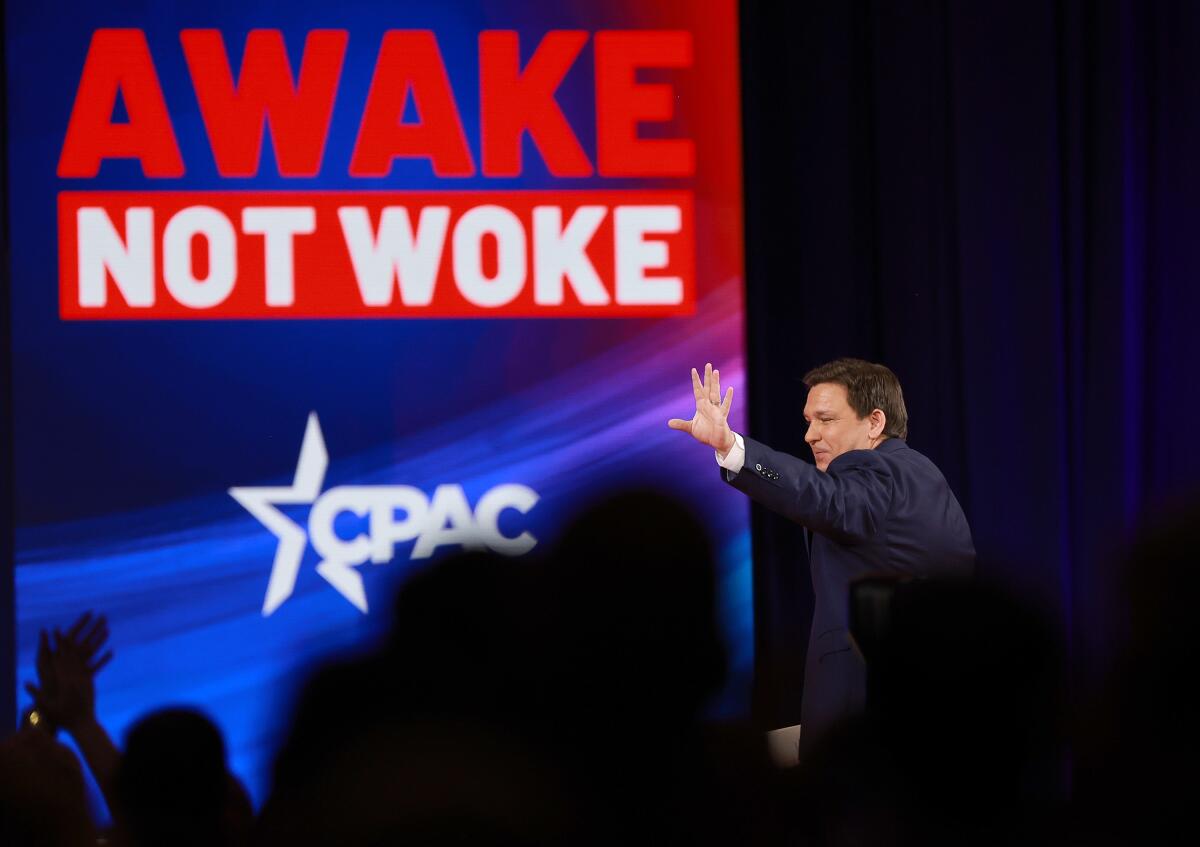 A man walks onto a stage and waves under the projected words "Awake not woke" and the CPAC logo