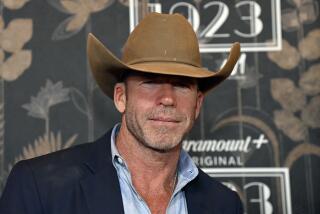 Taylor Sheridan smiles in a blue coat and shirt as a light brown cowboy hat casts a shadow on his eyes 