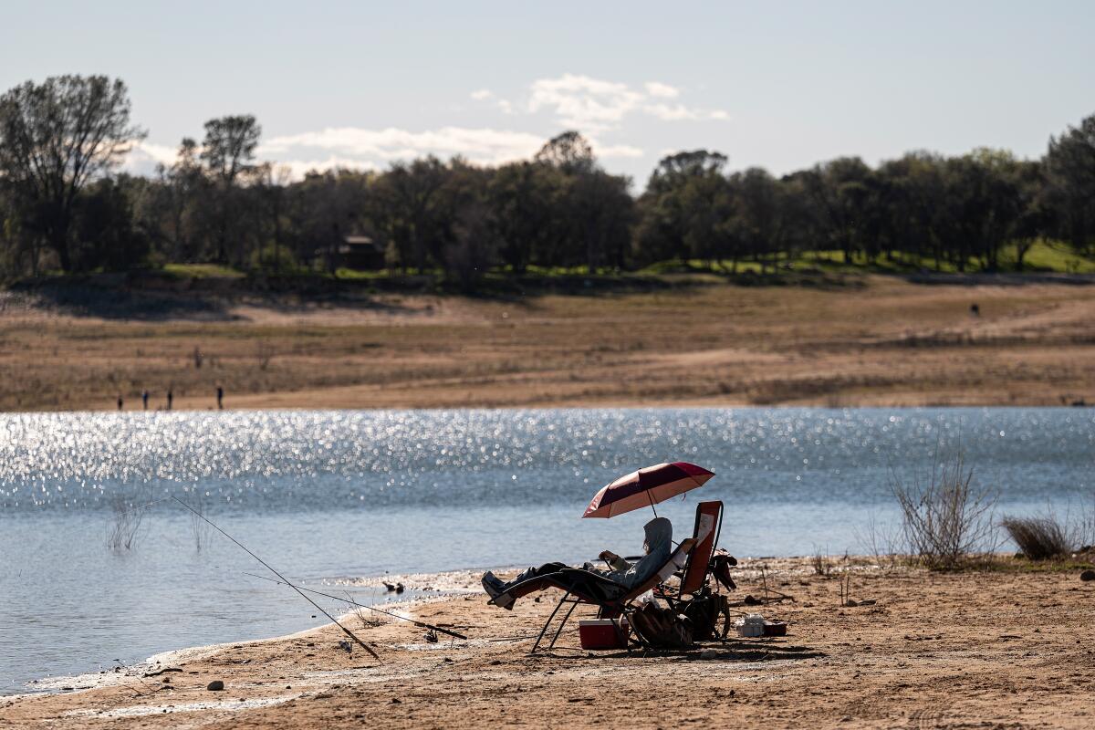 A person sits in a camp chair with an umbrella on a dry brown piece of land near the edge of a body of water.