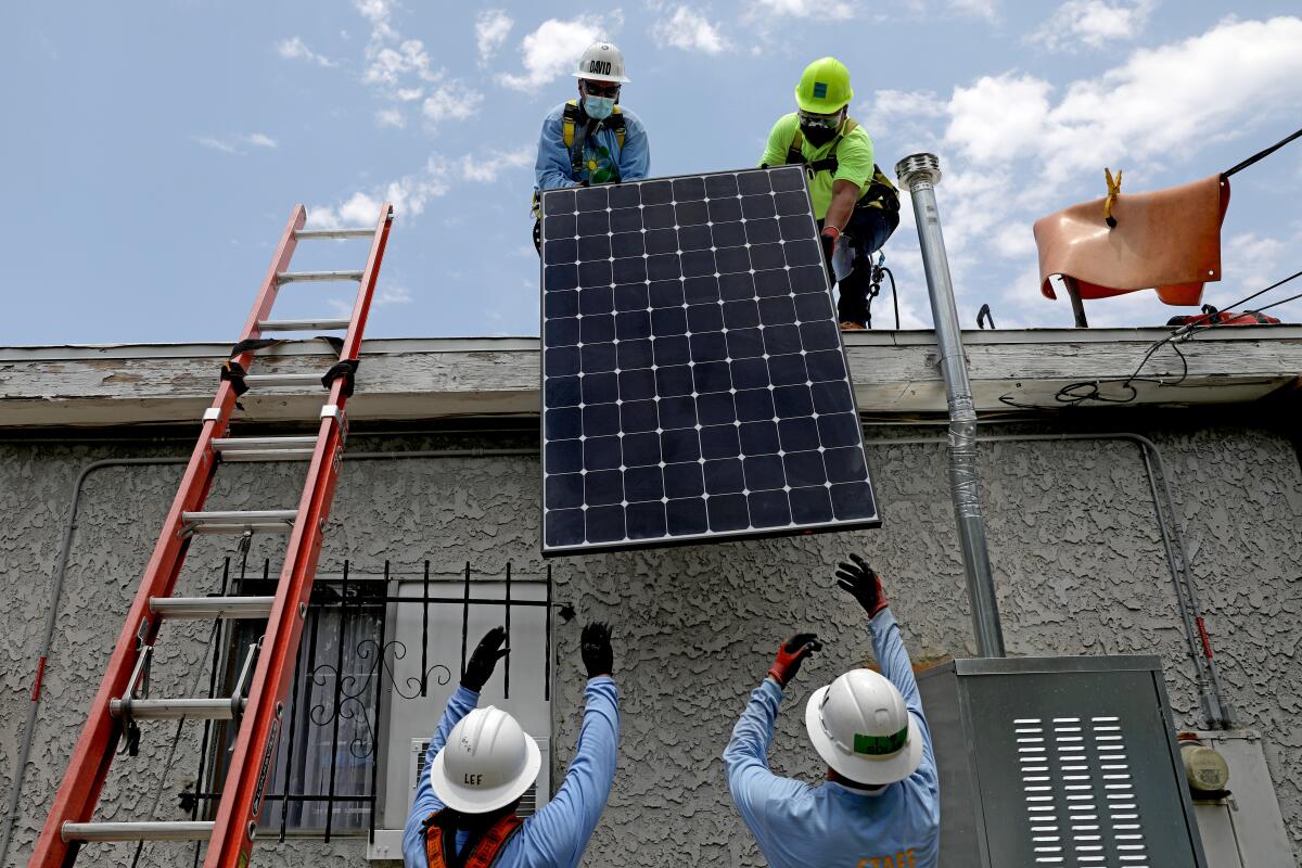 Two workers in hard hats hand a solar panel up to two other workers in hard hats on top of a house.